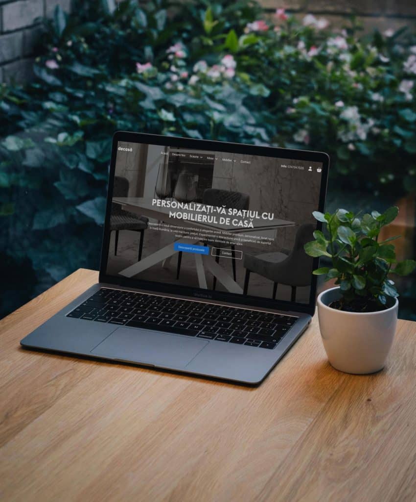Un laptop care afișează un site web de mobilier așezat pe o masă de lemn lângă o plantă mică în ghiveci, cu un perete de cărămidă și frunziș în fundal.