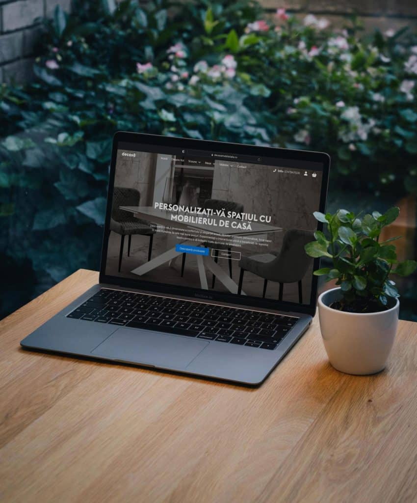 Un laptop pe o masă de lemn care afișează un site web despre personalizarea mobilierului de casă, lângă o plantă mică în ghiveci, cu un fundal de perete de cărămidă.